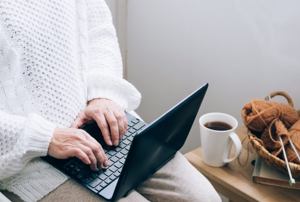 A middle-aged woman sits in a home chair with a mug of tea or coffee and works on a laptop.