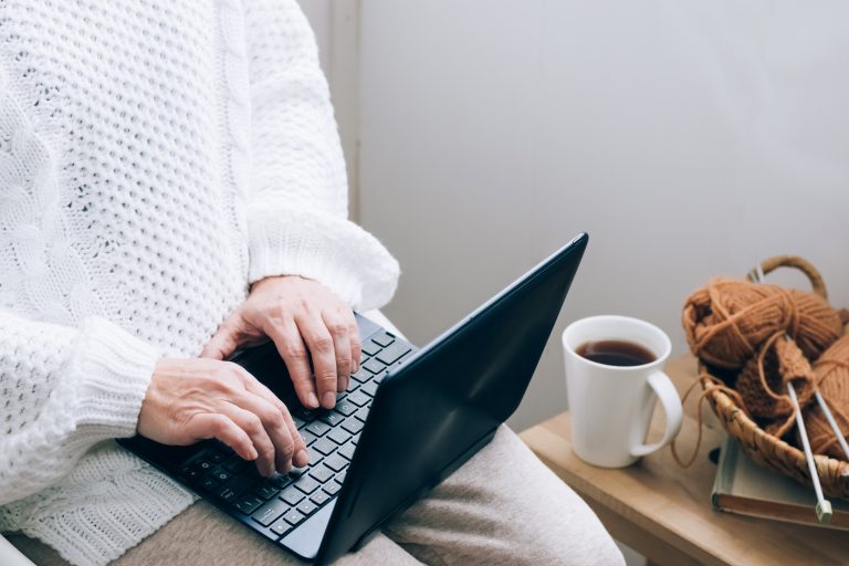 A middle-aged woman sits in a home chair with a mug of tea or coffee and works on a laptop.