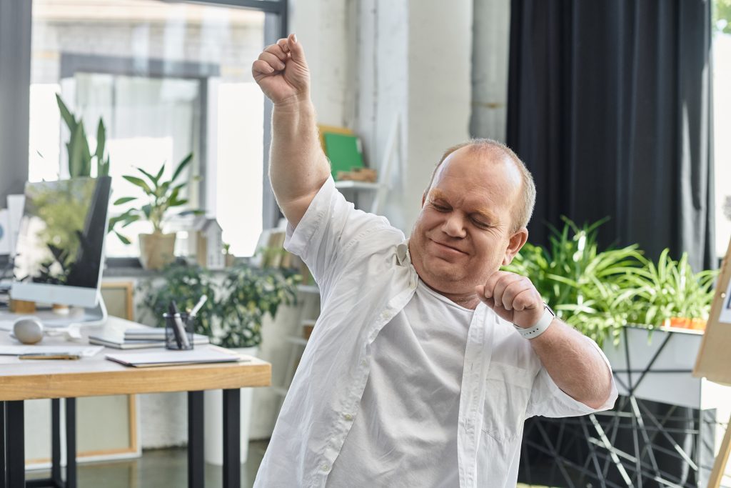 A man with inclusivity in a white shirt stretches his arms overhead while standing in an office setting.